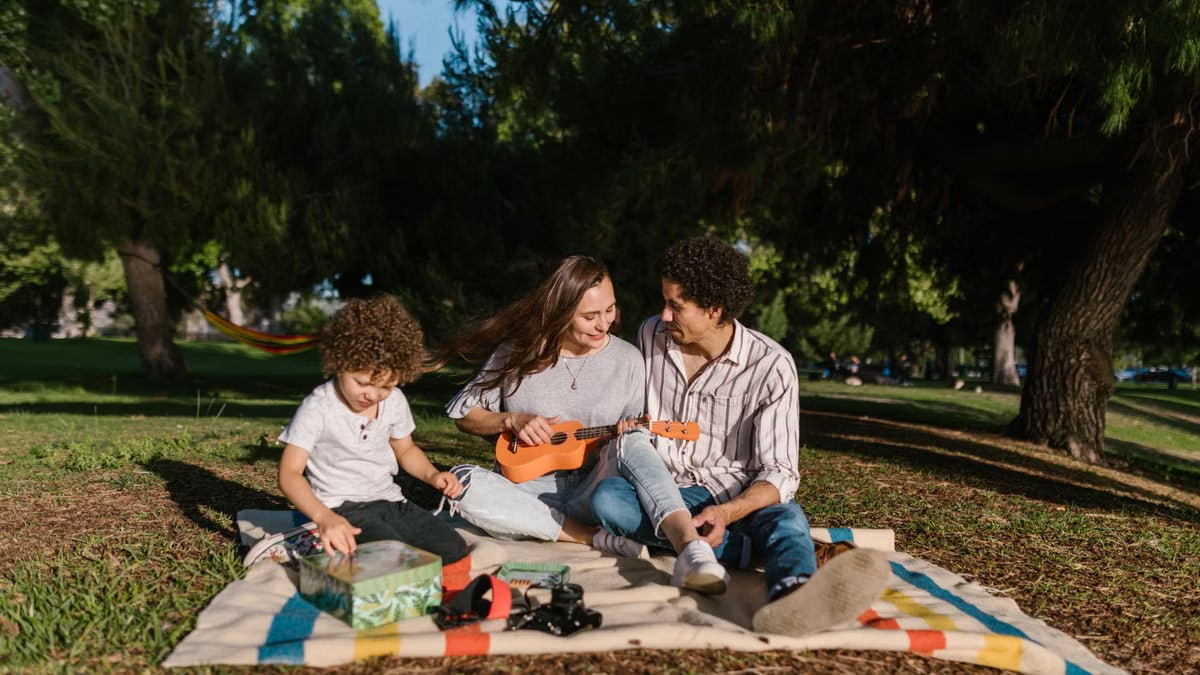 A joyful family enjoying a summer picnic with a ukulele in a sunny park setting.